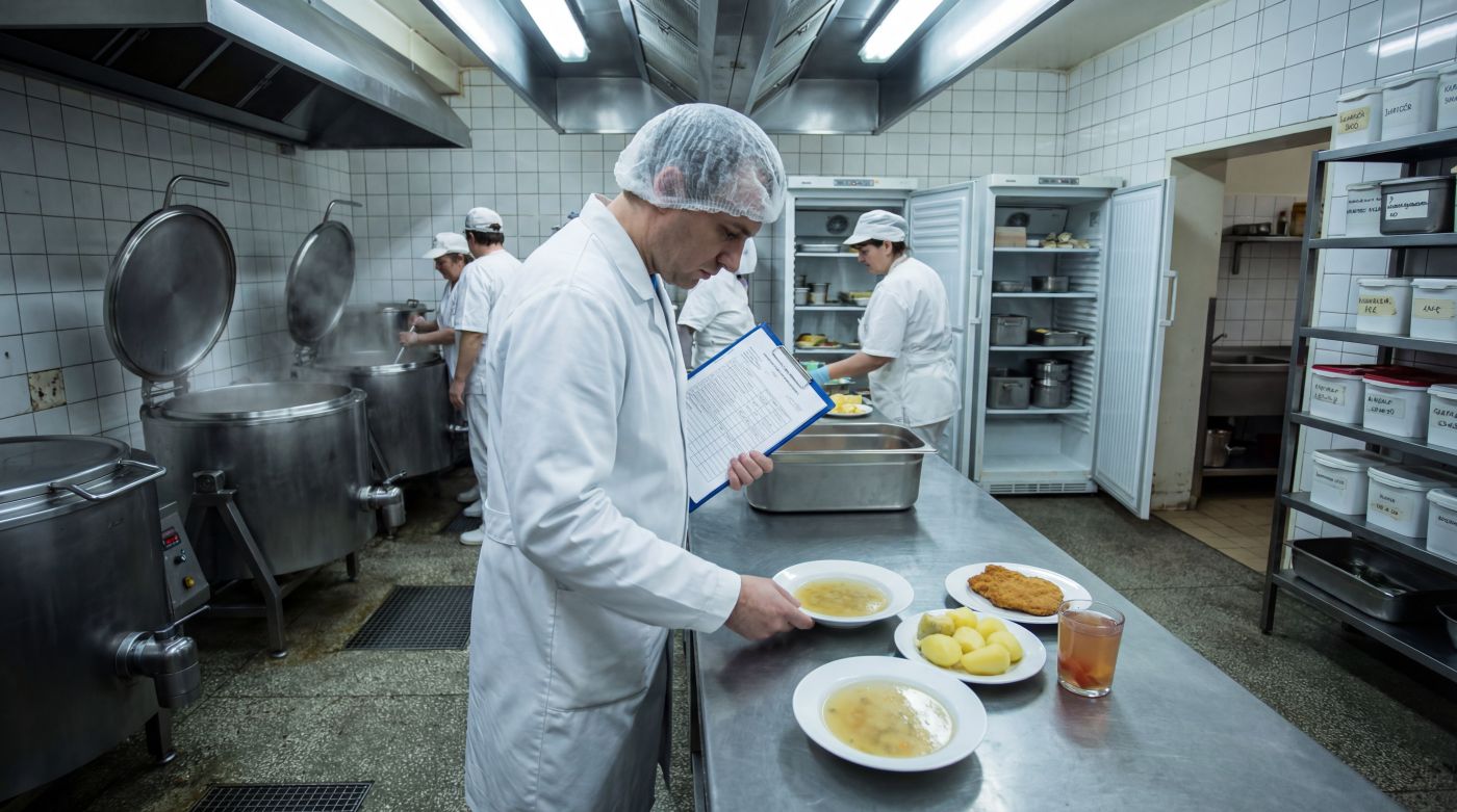 Pracownik w białym fartuchu i czepku w szpitalnej kuchni sprawdza talerze z zupą i obiadem, trzymając w ręku formularz kontrolny; w tle inni kucharze gotują w dużych kotłach, a lodówki i pojemniki z podpisami stoją otwarte, co ilustruje kontrolę sanepidu w szpitalu lub DPS-ie w latach 2023–2025.